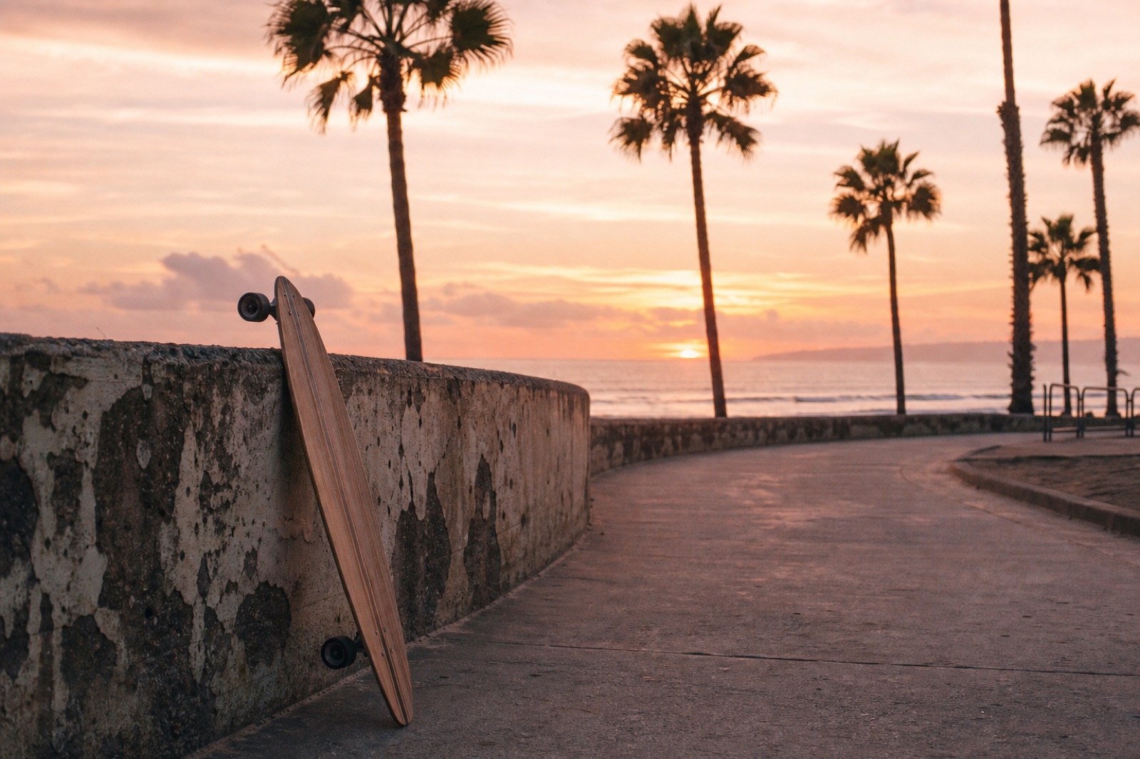 Sunrise over a California beach boardwalk, a longboard leaning against a weathered stone wall